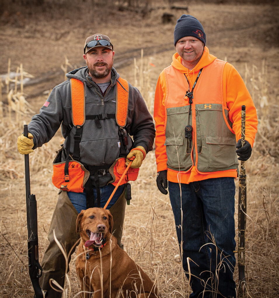 Two men dressed in hunter's orange holding shotguns, a dog sits next to them