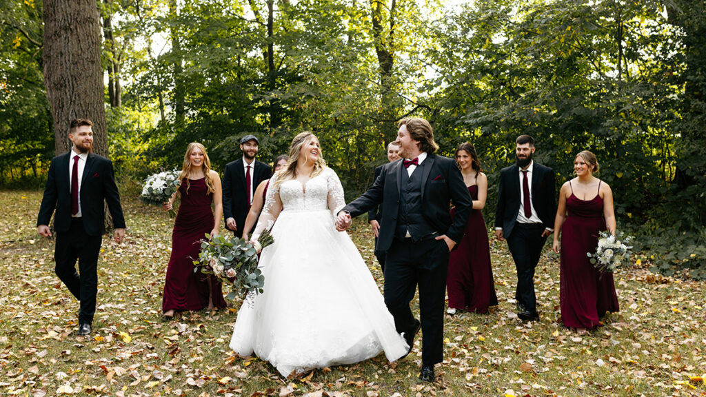 A newly wed couple with their wedding party behind them, walking through the woods