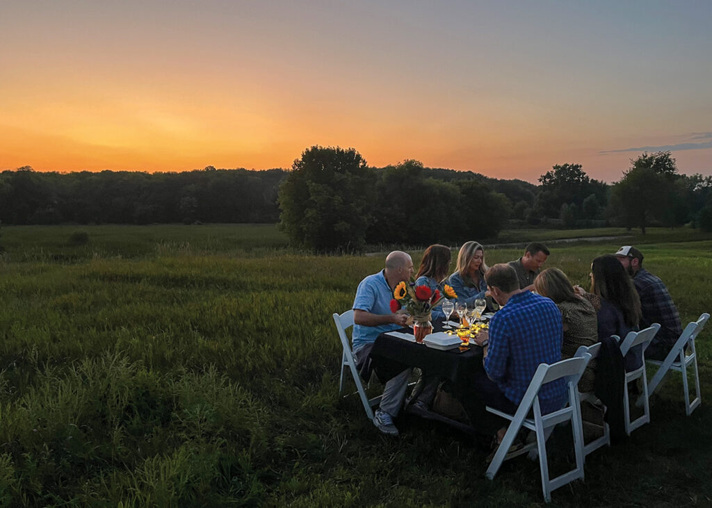 A small group gathered at a picnic table in an empty grass field as the sun sets behind them