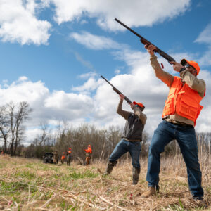 Men in upland gear taking aim at the sky with shotguns in a field.