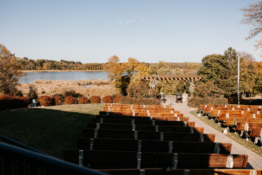 Wood arbor with flowers on sides. Pews leading up to the arbor. Water and tree view behind the arbor.
