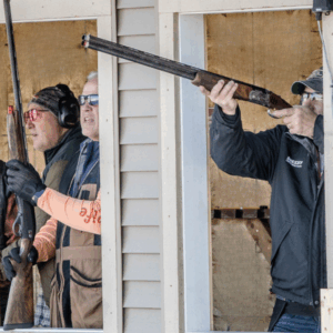 3 men looking out 5-stand windows, one ready to take shot at clay target with shotgun